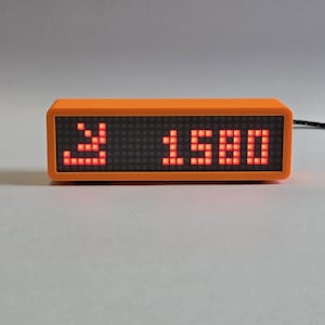 Op de afbeelding: Oranje rechthoekige digitale klok met het nummer "1580" in rode LED-lampjes. De klok toont ook een rood LED-symbool. Een zwart snoer is bevestigd.