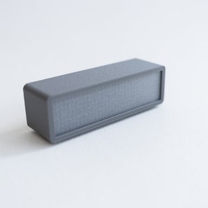Puede incluir: Un peque&ntilde;o altavoz rectangular gris con una superficie texturizada.