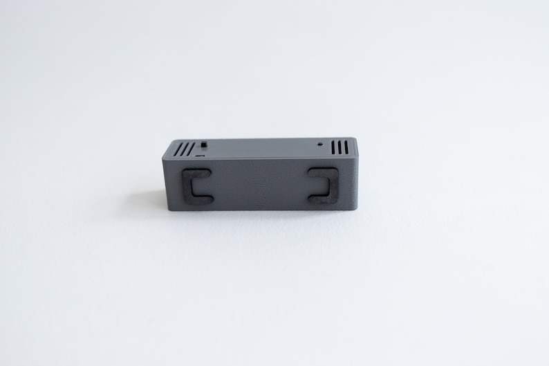 Puede incluir: Una caja rectangular gris con dos pies de goma negra en la parte inferior.