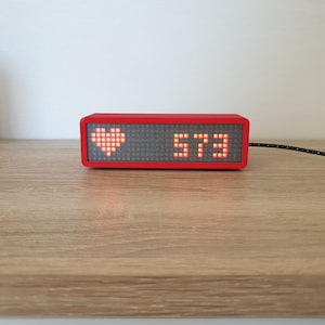 May include: A red rectangular device with a digital display showing the number 573 and a red heart symbol. The device is connected to a black and yellow cord.