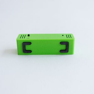 May include: A small, green rectangular box with two black, rectangular handles on the sides. The box has a textured surface and appears to be made of plastic.