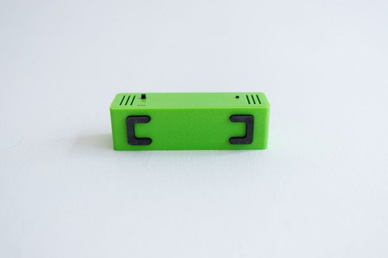 May include: A small, green rectangular box with two black, rectangular handles on the sides. The box has a textured surface and appears to be made of plastic.