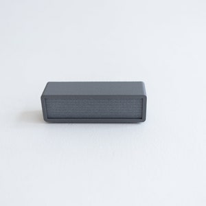Puede incluir: Un peque&ntilde;o altavoz rectangular gris con una rejilla de tela.