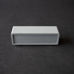 Puede incluir: Un peque&ntilde;o altavoz rectangular gris con una superficie texturizada.