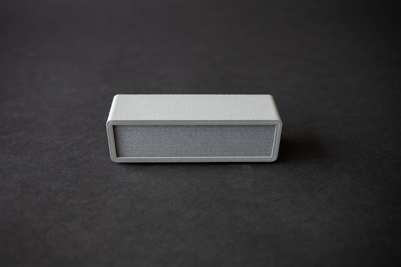 Puede incluir: Un peque&ntilde;o altavoz rectangular gris con una superficie texturizada.