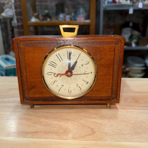 May include: A vintage wooden clock with a gold-tone face and hands. The clock has a rectangular wooden case with a handle on top and small brass feet. The clock face displays the numbers 1 through 12.