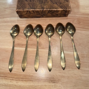 May include: Set of six vintage gold-toned spoons with long handles, arranged on a wooden surface. The spoons are in a row, with a small design on the handle. A decorative box with a brown patterned design is in the background.