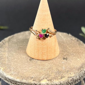 May include: A gold ring with three heart-shaped gemstones. The gemstones are green, pink, and black.