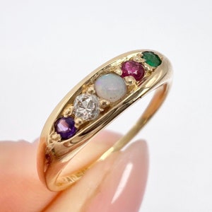 Op de afbeelding: Een gouden ring met vijf edelstenen. De stenen zijn een paarse amethist, een witte diamant, een witte opaal, een rode robijn en een groene smaragd.