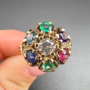 May include: A gold-toned ring featuring a central clear stone surrounded by a floral arrangement of colorful gemstones. The ring's design includes blue, green, purple, and pink stones, creating a vibrant and eye-catching piece of jewelry.