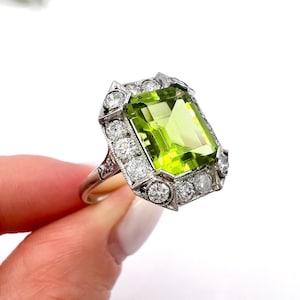 May include: A silver ring featuring a large, rectangular, light green gemstone surrounded by smaller, clear gemstones. The ring has an Art Deco style with intricate detailing around the main stone and smaller stones.