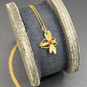 May include: A gold chain necklace with a bee pendant featuring a red gemstone. The bee is adorned with small white pearls.