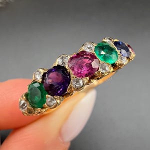 Op de afbeelding: Een gouden ring met een rij kleurrijke edelstenen: groen, paars, roze, groen en blauw. De ovale edelstenen zijn omgeven door kleine, heldere stenen. De ring wordt tegen een donkere achtergrond getoond.