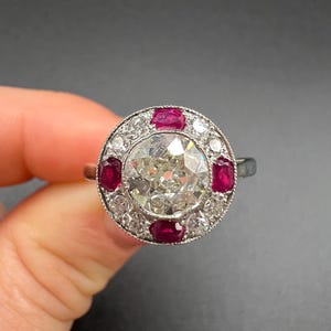 Könnte beinhalten: Ein silberner Ring mit einem großen, runden, klaren Diamanten in der Mitte, umgeben von kleineren, klaren Diamanten und sechs rechteckigen roten Steinen. Der Ring wird vor einem dunklen Hintergrund gehalten.