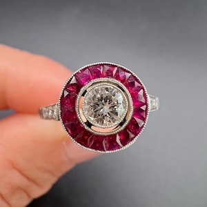 Op de afbeelding: Een zilverkleurige ring met een centrale ronde briljant geslepen diamant in een zetting, omringd door een halo van dieprode, vierkant geslepen robijnen. De metalen band heeft gedetailleerde milgrain-afwerking en kleine accentdiamanten.