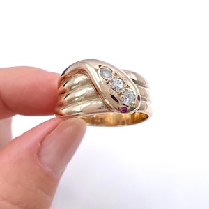 May include: A gold ring shaped like a coiled snake with a smooth, polished finish. The snake head features three clear, round-cut stones set in a row along the top, with two small, vibrant red stones serving as eyes at the very tip of the head.