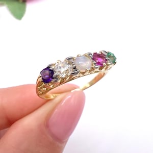 Può includere: Un anello in oro con una fila di cinque pietre preziose multicolori, tra cui un'ametista viola, una pietra bianca trasparente, un opale iridescente pallido, un rubino rosa vivace e un piccolo smeraldo verde, su una fascia in metallo decorata.