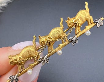 Op de afbeelding: Een goudkleurige broche met drie katten die een tak beklimmen. De katten zijn verbonden door kettingen en versierd met kleine parels. De tak heeft zilverkleurige bladdetails.