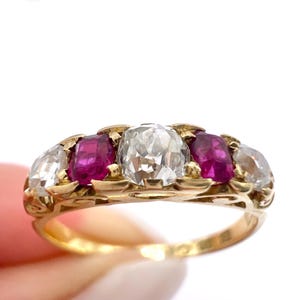 May include: A gold ring with a central clear diamond, two red gemstones, and two clear diamonds. The ring has an ornate, filigree-style band. The gemstones are set in a gold band.