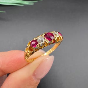 May include: A gold ring featuring a row of alternating round-cut red rubies and clear white diamonds. The band has an intricate, openwork filigree design along the sides, showcasing a repeating pattern of metal loops that hold the five gemstones.
