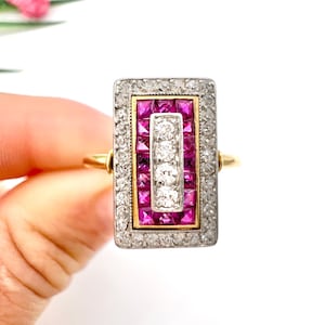 May include: A rectangular ring with a gold band and a silver-colored top. The top features a central row of white stones flanked by rows of pink stones, all surrounded by smaller white stones. The ring is held by a hand.