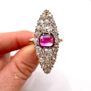 May include: A gold ring with a central magenta gemstone, rectangular in shape, encircled by smaller, clear gemstones. The ring's design is elongated and teardrop-shaped, with the gemstones arranged in a clustered formation.