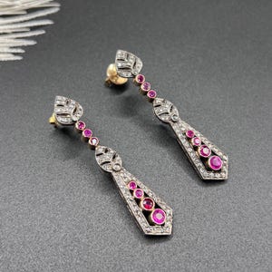 May include: A pair of drop earrings featuring a leaf-shaped top section, a slender stem with small round pink stones, and a larger geometric base adorned with multiple round pink stones and small clear stones set in a dark metal frame.