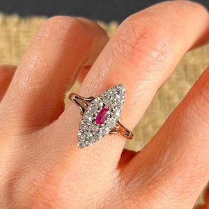 May include: A gold ring featuring a red gemstone surrounded by smaller white gemstones in a marquise shape.