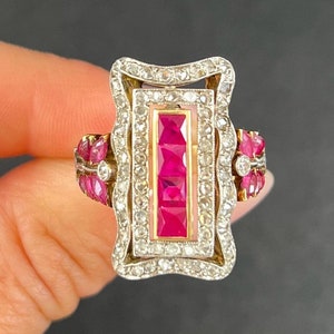 Puede incluir: Un anillo vintage con una piedra de rubí rectangular rodeada de diamantes. El anillo está engastado en una banda de metal blanco y tiene un diseño decorativo.