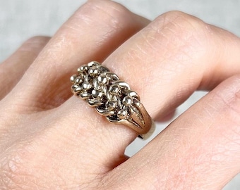 Könnte beinhalten: Ein goldfarbener Ring mit einem Cluster aus kleinen, strukturierten Perlen. Der Ring wird an einem Finger getragen und hat ein detailliertes Design. Das Band des Rings ist ebenfalls goldfarben, und der Stil ist Vintage oder antik.