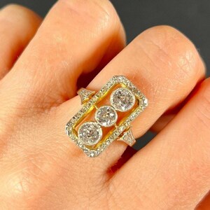 Puede incluir: Un anillo de oro con un engaste rectangular que presenta tres diamantes redondos. Los diamantes están rodeados de diamantes más pequeños.