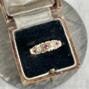 Könnte beinhalten: Ein goldener Ring mit roten und weißen Edelsteinen, die in einer Bandfassung gefasst sind. Der Ring befindet sich in einer kleinen braunen Schachtel mit dem Text "W.A. Muir Ltd. Jewellers & Diamond Merchants 8, Market Place, 45, Stockport Road, 25, Upper Jackson St, Manchester" auf der Innenseite des Deckels.