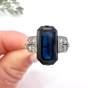 May include: A vintage ring featuring a large, rectangular, dark blue gemstone set in a silver-toned metal. The ring has smaller, clear gemstones on either side of the main stone, adding sparkle. The design is classic and elegant.