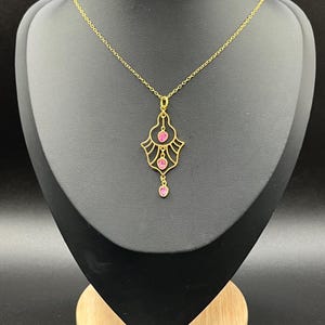 Könnte beinhalten: Eine goldfarbene Halskette mit einem Anhänger mit rosa Edelsteinen. Der Anhänger hat ein verziertes Design mit drei herzförmigen Steinen und einem herabhängenden tropfenförmigen Stein. Die Halskette wird auf einer schwarzen Schmuckbüste präsentiert.