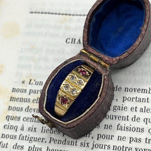 May include: A gold ring featuring two red gemstones and two clear stones set in a row, housed in an oval-shaped, hinged jewelry box with a dark brown exterior and a vibrant blue velvet interior lining, resting on a page of vintage printed text.