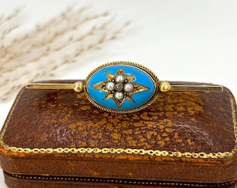 May include: An antique gold-tone brooch with a central turquoise-colored oval, featuring a starburst design with small pearls. The brooch is set on a brown leather jewelry box with gold trim. A classic piece of jewelry.