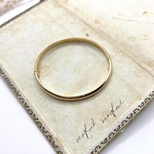 May include: A gold-coloured bangle bracelet with a simple, rounded design. The bracelet is resting on an open, antique book with handwritten text and decorative borders. The word "useful" is visible on the page.