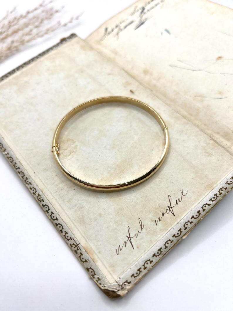 May include: A gold-coloured bangle bracelet with a simple, rounded design. The bracelet is resting on an open, antique book with handwritten text and decorative borders. The word "useful" is visible on the page.