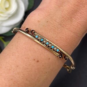 Pode incluir: Uma pulseira rígida de tom dourado com uma barra horizontal central adornada com uma fila de pequenas pedras redondas que alternam entre turquesa e branco perolado. A barra apresenta ornamentos de volutas em cada extremidade do metal.