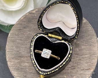 May include: A gold ring with a square-cut clear gemstone set in a prong setting, displayed inside a black heart-shaped jewellery box with gold-coloured trim and a black velvet interior lining, featuring a hinged lid and a white fabric interior.