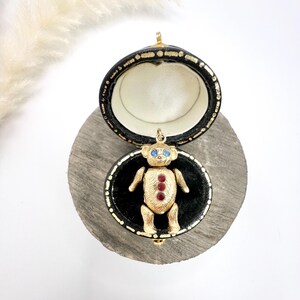 Op de afbeelding: Een gouden teddybeerbedel met blauwe ogen en rode edelsteenaccenten, gepresenteerd in een zwarte doos met een fluwelen voering.