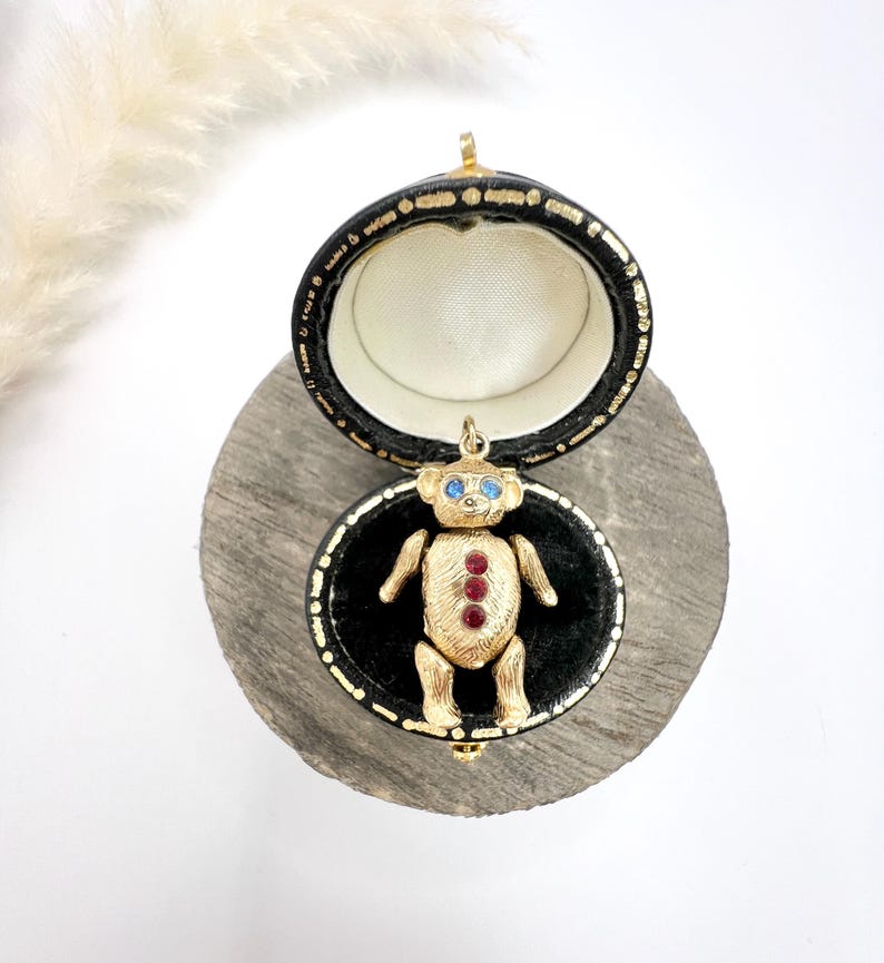 Op de afbeelding: Een gouden teddybeerbedel met blauwe ogen en rode edelsteenaccenten, gepresenteerd in een zwarte doos met een fluwelen voering.