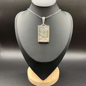 Puede incluir: Un collar con colgante de plata con un colgante rectangular. El colgante tiene un diseño de sello con una escena de naturaleza detallada, un marco plateado y una cadena delicada. La escena incluye pájaros y plantas.