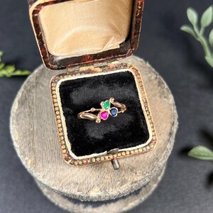 May include: A gold ring with three heart-shaped gemstones: green, pink, and blue. The ring is in a small, brown velvet box.