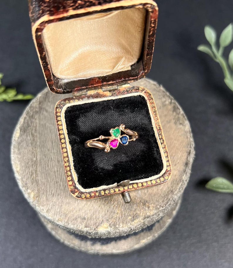 May include: A gold ring with three heart-shaped gemstones: green, pink, and blue. The ring is in a small, brown velvet box.