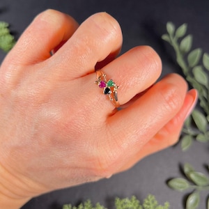 May include: A gold ring with three heart-shaped gemstones: black, green, and pink.