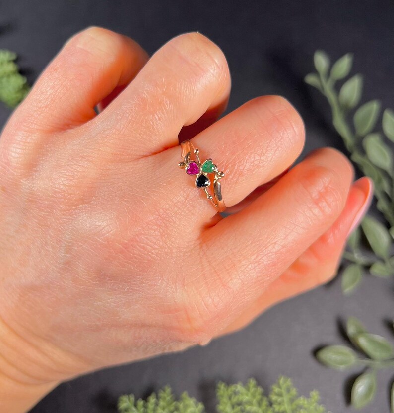 May include: A gold ring with three heart-shaped gemstones: black, green, and pink.