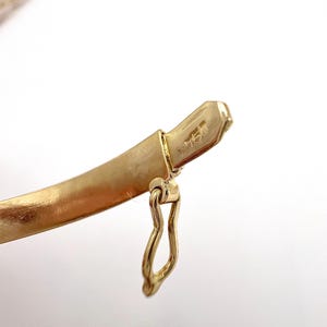 May include: Close-up of a gold bracelet clasp. The clasp is a curved, solid band with a small, attached hook. The metal has a matte finish and a hallmark stamp. The background is white.