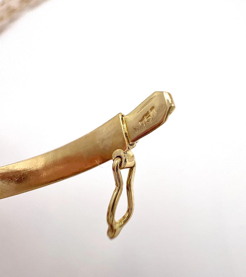 May include: Close-up of a gold bracelet clasp. The clasp is a curved, solid band with a small, attached hook. The metal has a matte finish and a hallmark stamp. The background is white.