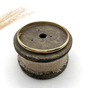 May include: A gold bangle bracelet with a simple design. The circular bracelet is made of metal and sits on a wooden spool with a beige fabric wrap. The background is white. The bracelet's diameter is approximately 6.5 cm.